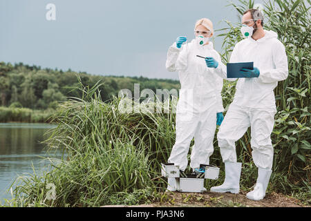 Homme libre de masque et écrit dans le presse-papiers et de pointage pendant que son collègue de l'échantillon de l'examen de l'eau dans la fiole de test pis Banque D'Images