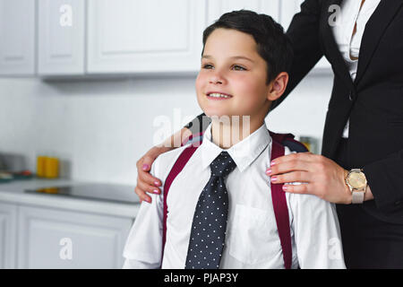 Vue partielle de mère en costume et fils dans l'uniforme scolaire avec sac à dos dans la cuisine à la maison, concept retour à l'école Banque D'Images