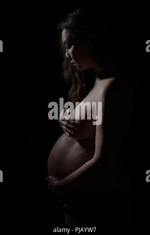 Un pregnant Asian woman posing sur le côté, regardant vers le bas. Un sombre portrait noir et blanc artistique. Banque D'Images