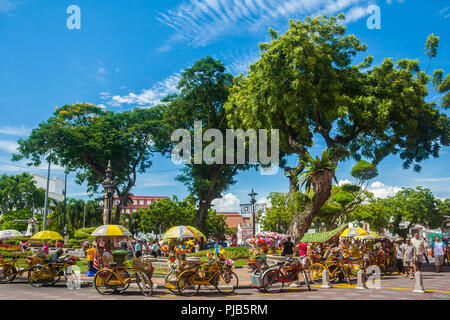 Les touristes se promener le long de l'joliment décorées trishaws jaune près de la fontaine à la Dutch Square (zone Stadthuys) sous les arbres de l'ombre sur une belle... Banque D'Images