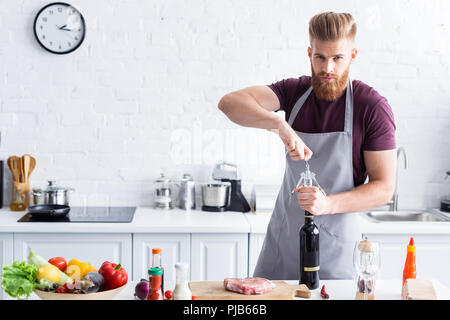 Beau jeune homme barbu tablier en ouvrant une bouteille de vin et à la caméra au moment de la cuisson dans la cuisine Banque D'Images