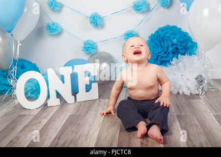 Portrait de pleurer crier malheureux Caucasian baby boy en pantalon foncé sur son premier anniversaire avec des lettres et des ballons, l'un assis sur un plancher en bois je Banque D'Images