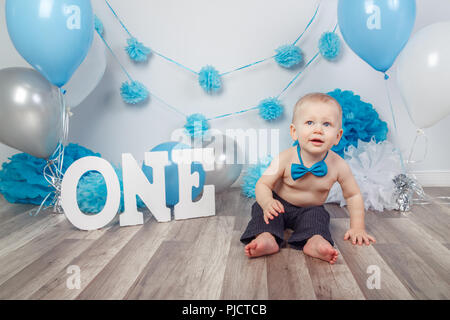 Portrait de l'adorable petit garçon de race blanche dans un pantalon foncé et bleu noeud papillon fêter son premier anniversaire avec l'un des lettres et des ballons, assis sur woode Banque D'Images