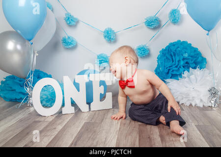 Portrait de pleurer crier malheureux Caucasian baby boy en pantalon foncé et rouge noeud papillon sur son premier anniversaire avec des lettres et des ballons, l'un assis o Banque D'Images