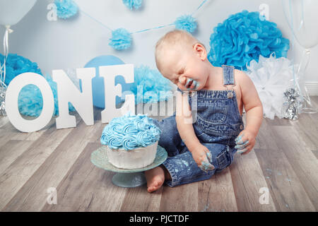 Portrait of cute adorable pleurer triste Caucasian baby boy en jeans dans l'ensemble célébrer son premier anniversaire avec un gâteau gourmand et lettres ballons cak Banque D'Images