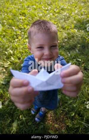 Maintenez l'enfant rire heureux navire papier à l'intérieur. Champ vert arrière-plan. Enfance heureuse, été, vacances, voyages, vacances Banque D'Images