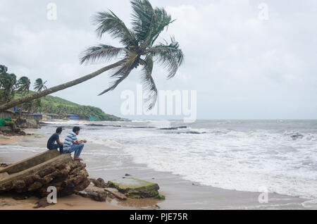 Deux garçons assis sous un cocotier, regardant dans le fracas des vagues, Inde Banque D'Images