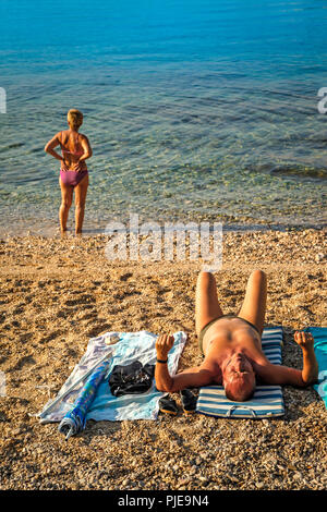 Une femme au bord de l'eau fixe sur l'Adriatique sur l'île croate de Krk au resort de Baska, tandis que l'homme des bains de soleil sur la plage Banque D'Images