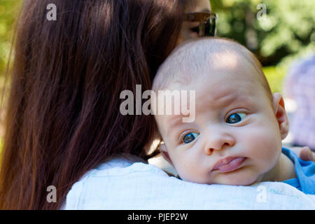 Portrait de beau bébé aux yeux bleus et mignon visage, la mère avec les cheveux bruns, c'est servir bébé sur son épaule, des rots bébé, position à l'extérieur de jour Banque D'Images
