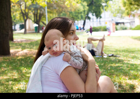 Mère et bébé dans le parc public, mère amoureuse compassion cute infant dans les mains de faire un rot après la tétée, le menton de bébé repose sur l'épaule de maman Banque D'Images