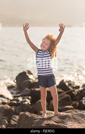 Petite fille aux mains levées avec sur un rocher près d'une mer Banque D'Images