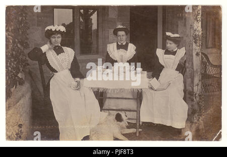 Carte postale originale, claire et édouardienne de femmes de ménage habilement habillées, portant des tabliers et des casquettes, prenant une pause-thé, à l'extérieur dans le jardin d'une maison de campagne, Bridgwater, Somerset, Royaume-Uni vers 1905 Banque D'Images