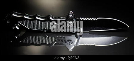 Black Ops tac couteau avec lame noire dentelée et coupe-ceinture de sécurité réfléchissant sur verre noir. Grade militaire printemps aider couteau. Banque D'Images