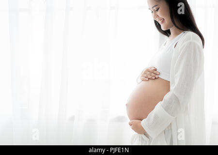 Close up of happy smiling pregnant ASian woman standingand de toucher son ventre à la maison. La grossesse, le repos, les gens et l'attente concept Banque D'Images