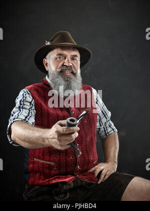 Portrait dramatique de barbu gris smiling senior man in hat fumeurs pipe. Vue sur la tyrolienne, autrichienne, vieil homme bavarois en costume traditionnel national en style rétro. Banque D'Images