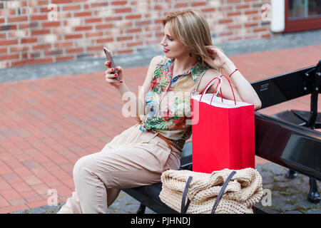 Vue latérale du profil belle jeune femme vêtue de chemise tropicale et l'emplacement sur banc noir dans la rue à la recherche et le smart phone après shop Banque D'Images