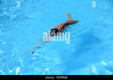 Man en nage libre dans une piscine extérieure sur une journée ensoleillée. L'homme porte un maillot rouge et un capuchon noir. fit man natation pendant les loisirs Banque D'Images