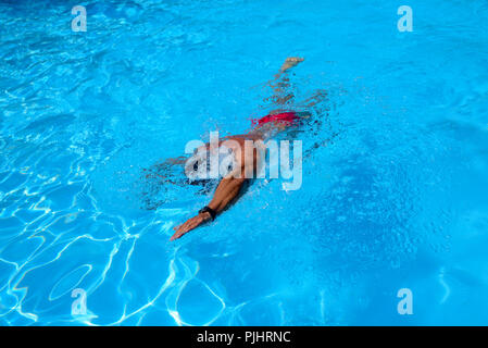 Man en nage libre dans une piscine extérieure sur une journée ensoleillée. L'homme porte un maillot rouge et un capuchon noir. fit man natation pendant les loisirs Banque D'Images