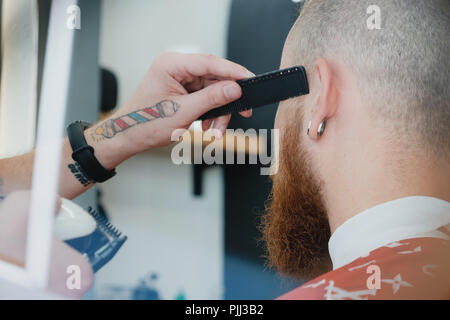 Un beau jeune homme barbu dans un salon de coiffure. Le salon de coiffure brosses ses cheveux et coupe avec une tondeuse électrique. Banque D'Images