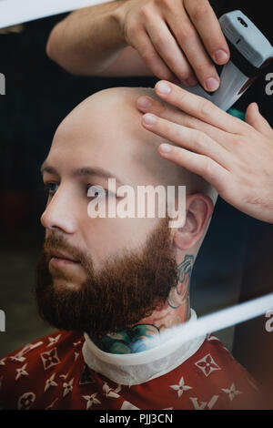 Un bel homme barbu dans un skinhead barbershop. Le barbier se rase la tête avec une tondeuse électrique. Banque D'Images