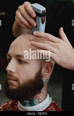 Un bel homme barbu dans un skinhead barbershop. Le barbier se rase la tête avec une tondeuse électrique. Banque D'Images