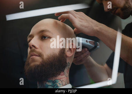 Un bel homme barbu dans un skinhead barbershop. Le barbier se rase la tête avec une tondeuse électrique. Banque D'Images