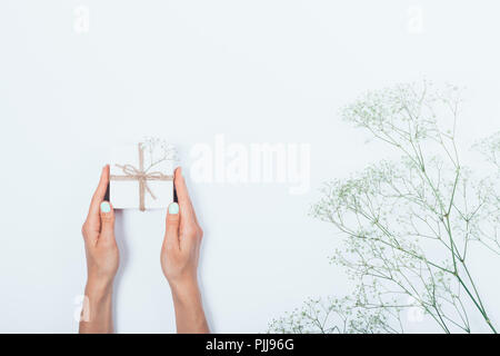 Woman's hands holding petit cadeau fort lié avec une corde près de gypsophila fleurs sur fond blanc, vue du dessus. Mise à plat de la composition minimale donnant une pr Banque D'Images