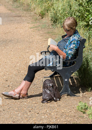 Femme d'âge moyen assis sur un banc, lisant un livre sur une belle journée d'été. dame plus âgée se détendre dans le soleil avec un bon livre à lire. Banque D'Images