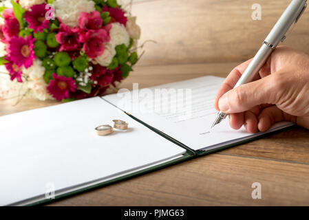 L'homme de signer un registre de mariage comme un époux ou un témoin avec un élégant stylo plume avec deux anneaux de mariage et les fleurs rouges, symbolique de l'amour. Banque D'Images