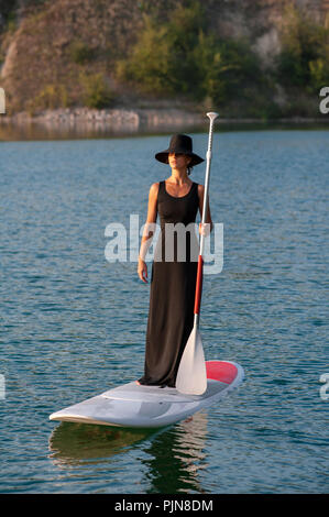 Silhouette d'une belle femme dans une robe noire sur stand up paddle board, le coucher du soleil. SUP Banque D'Images