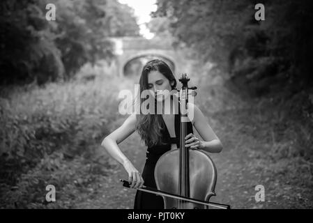Une jeune femme blonde joue du violoncelle à l'extérieur, derrière elle, sur un chemin d'un pont peut être vu Banque D'Images