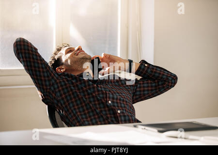 Relaxant du dos de l'entrepreneur sur sa chaise et on cell phone assis à son bureau. L'homme de prendre une pause du travail et talking on mobile phone leanin Banque D'Images