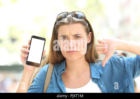 Vue avant, portrait d'une fille en colère montrant l'écran du téléphone en blanc avec le pouce vers le bas dans la rue Banque D'Images