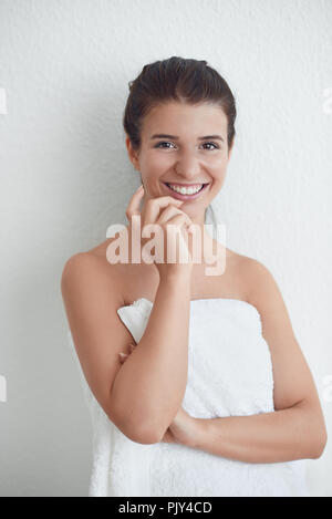 Heureux sain sourire séduisant jeune femme enveloppée dans une serviette blanche pelucheuse propre debout contre un mur blanc de l'hygiène personnelle et conceptuel sp Banque D'Images