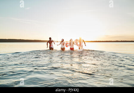 Coquille d'un groupe diversifié de jeunes amis en maillot de bain dans le milieu d'un lac au coucher du soleil, des éclaboussures et des vagues Banque D'Images