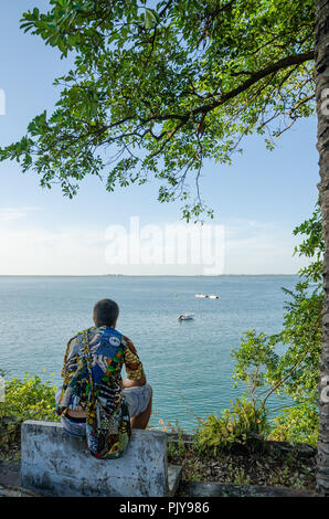 Vue arrière du jeune homme assis à l'autre donnant sur l'océan, portant chemise africaine colorée, Bubaque, îles Bijagos, Guinée Bissau Banque D'Images