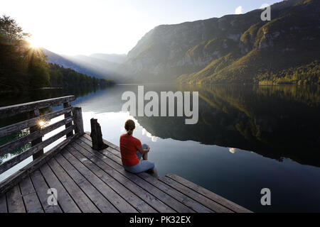 Femme assise sur une jetée en bois et à la recherche dans le coucher du soleil sur les montagnes, le lac de Bohinj, en Slovénie. Banque D'Images