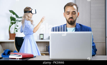 L'homme indépendant qui travaille à la maison sur l'ordinateur tandis que sa femme est de jouer à des jeux vidéo sur le casque de réalité virtuelle Banque D'Images