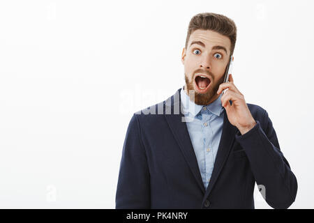 Wow mon entreprise a soulevé un autre million de dollars. Heureux et surpris beau mâle mature aux yeux bleus et à la barbe en costume officiel holding smartphone près de l'apprentissage de l'oreille, laissant tomber les détails choquants jaw surprise Banque D'Images