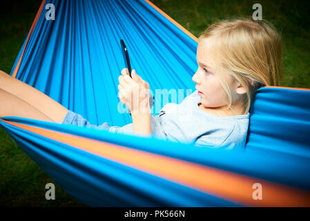 Enfant fille blonde jouant avec un téléphone intelligent en position allongée sur un hamac à l'extérieur Banque D'Images