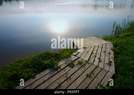 Vue panoramique du débarcadère de bateaux en bois par le lac au coucher du soleil avec la réflexion de la lumière. Banque D'Images