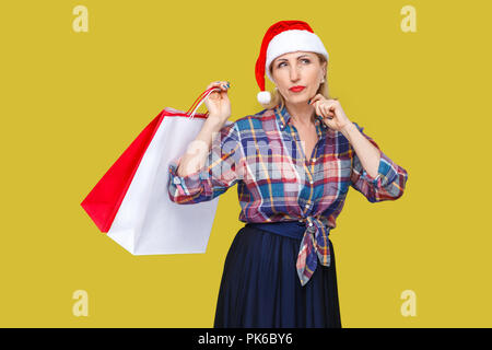 Portrait de femme d'âge moyen moderne méditant dans rouge santa cap et chemise à carreaux, permanent holding shopping bags avec visage, à la recherche en c Banque D'Images