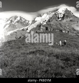 Années 1950, historiques, deux femmes marchant dans l'herbe des colline au-dessous des monts enneigés des montagnes suisses. Banque D'Images