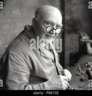 Années 1950, historique, un homme âgé dans un workcoat artisan traditionnel ou une salopette assis à son workbench la réparation outillage d'atelier, England, UK. Banque D'Images