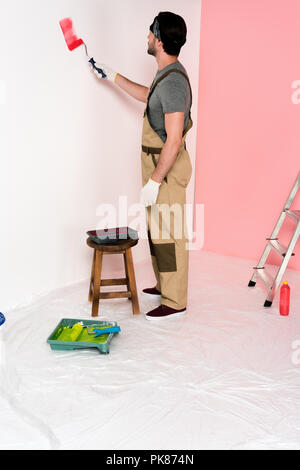 Jeune homme dans l'ensemble et de travail peinture bandeau rouge en mur par rouleau à peindre près de chaise avec plateau à rouleaux Banque D'Images