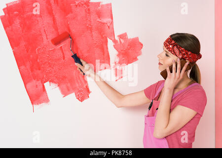 Vue latérale du jeune femme en bandeau et parler ensemble de travail sur smartphone et mur peinture en rouge par rouleau à peindre Banque D'Images