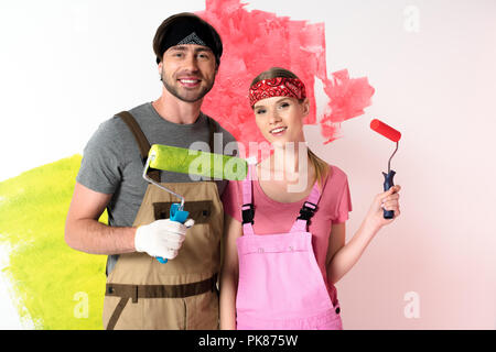 Jeune couple travaillant dans des combinaisons et bandeaux debout avec rouleaux à peinture en face de mur peint Banque D'Images