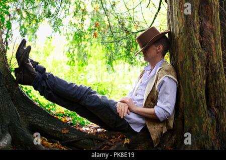 Un homme habillé comme un cowboy repose contre un tronc d'arbre Banque D'Images