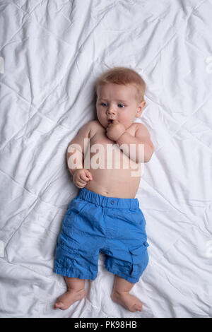 Satisfait peu réfléchies dans plus de bébé garçon pantalon bleu taille allongé sur le dos de sucer un pouce contre literie blanche vue d'en haut Banque D'Images