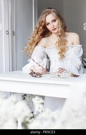 Girl écrit une lettre à son bien-aimé l'homme assis à la maison à la table dans une robe de lumière blanche, la pureté et l'innocence. Curly blonde romantique, bel Banque D'Images